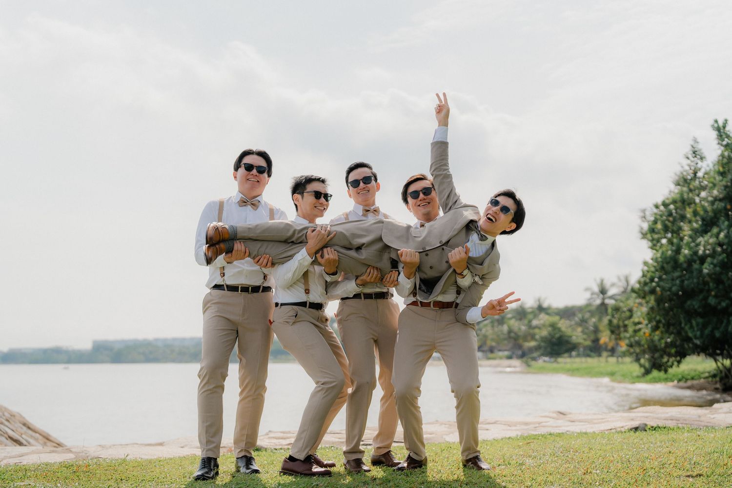 Groomsmen in khaki suits pose playfully outdoors by a waterfront on a sunny day.