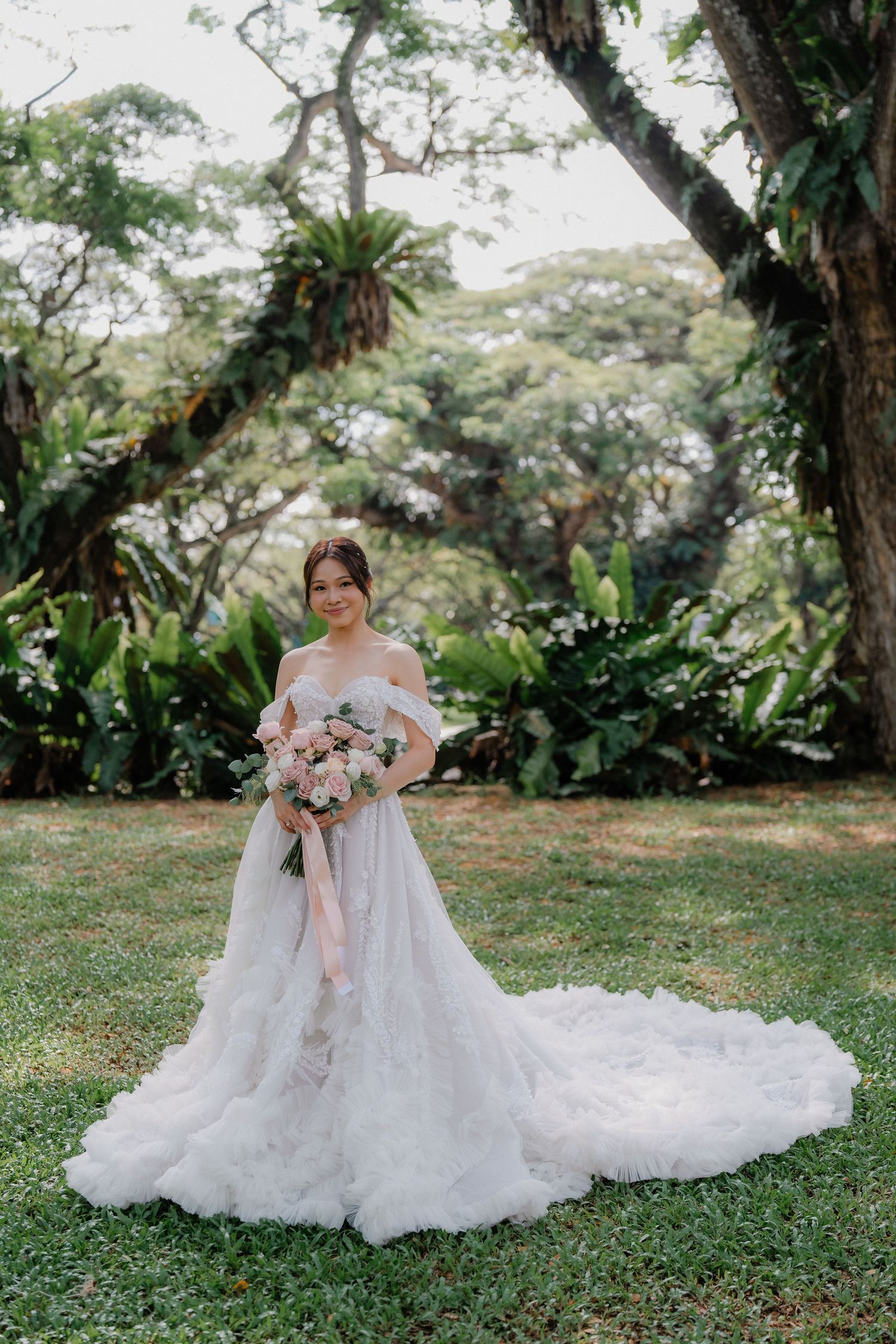 A bride in an elegant off-shoulder wedding gown with flowing train poses in a lush outdoor garden setting.