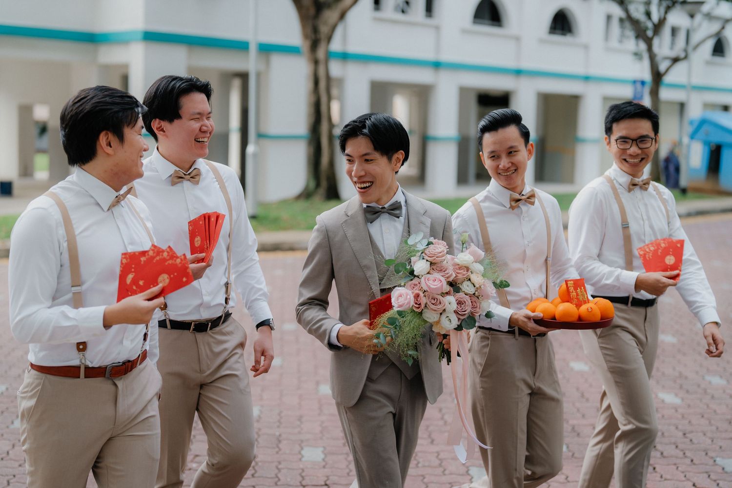 Groomsmen in white shirts and khaki pants walk together carrying red packets and floral bouquet.