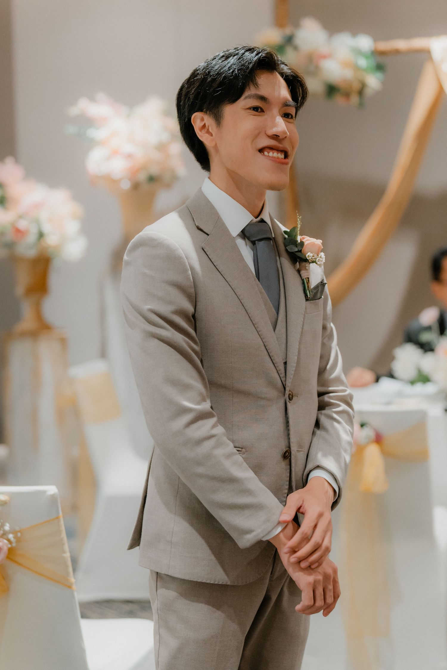 Groom wearing light grey suit and boutonniere smiles during wedding ceremony with floral decorations in background.