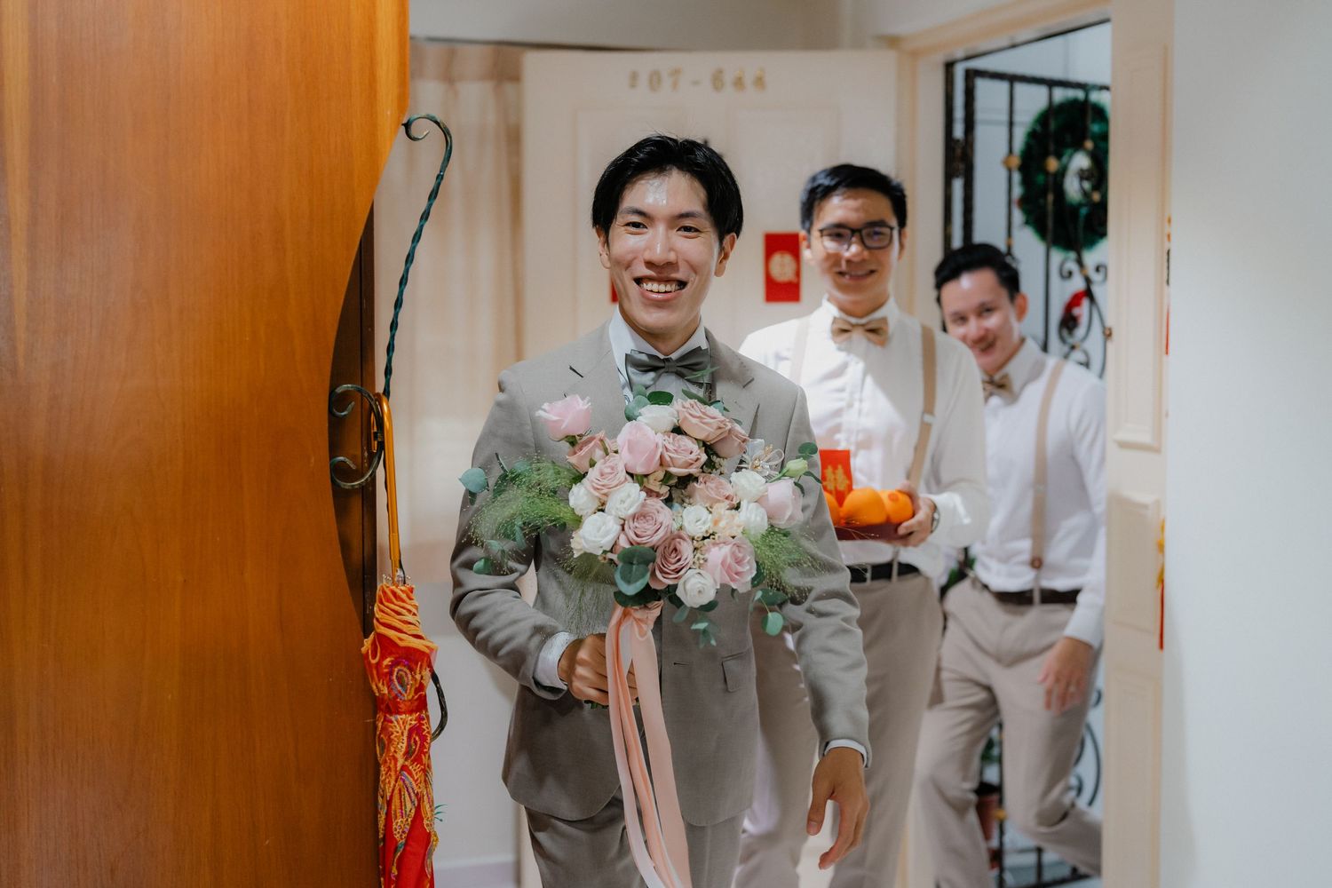 Groomsmen in light gray suits holding pink floral bouquets in a hallway.