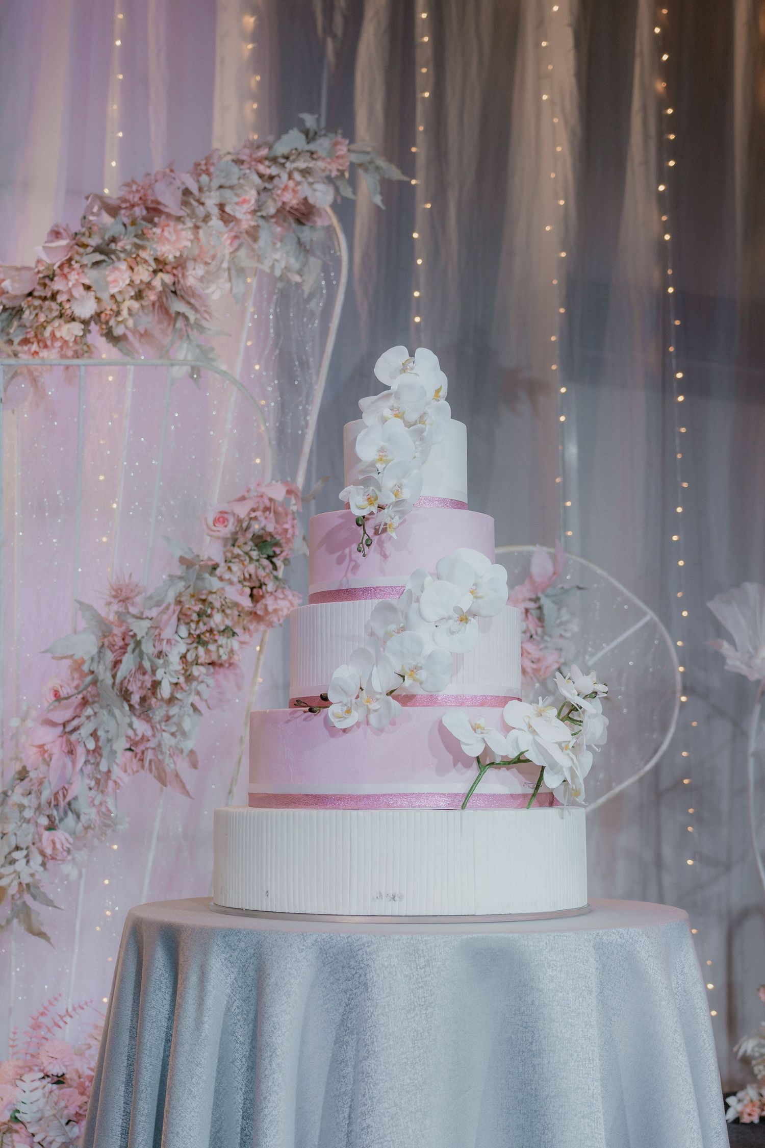 An elegant white wedding cake decorated with pink ribbons and cherry blossoms against a draped backdrop.