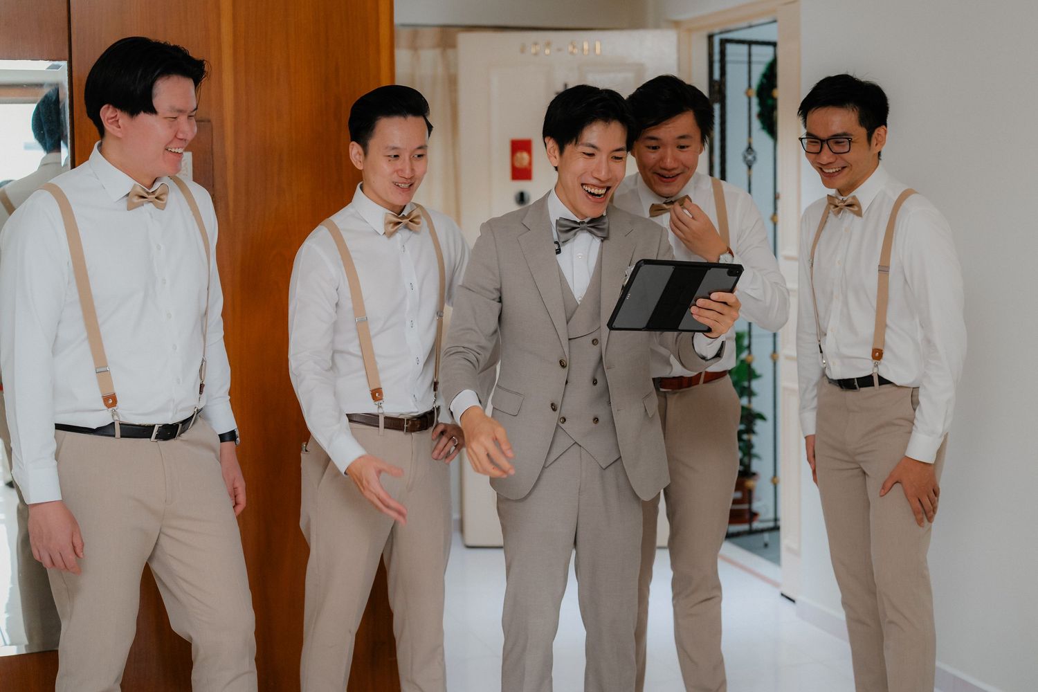 Groomsmen in light-colored suits and suspenders gather around a tablet device.