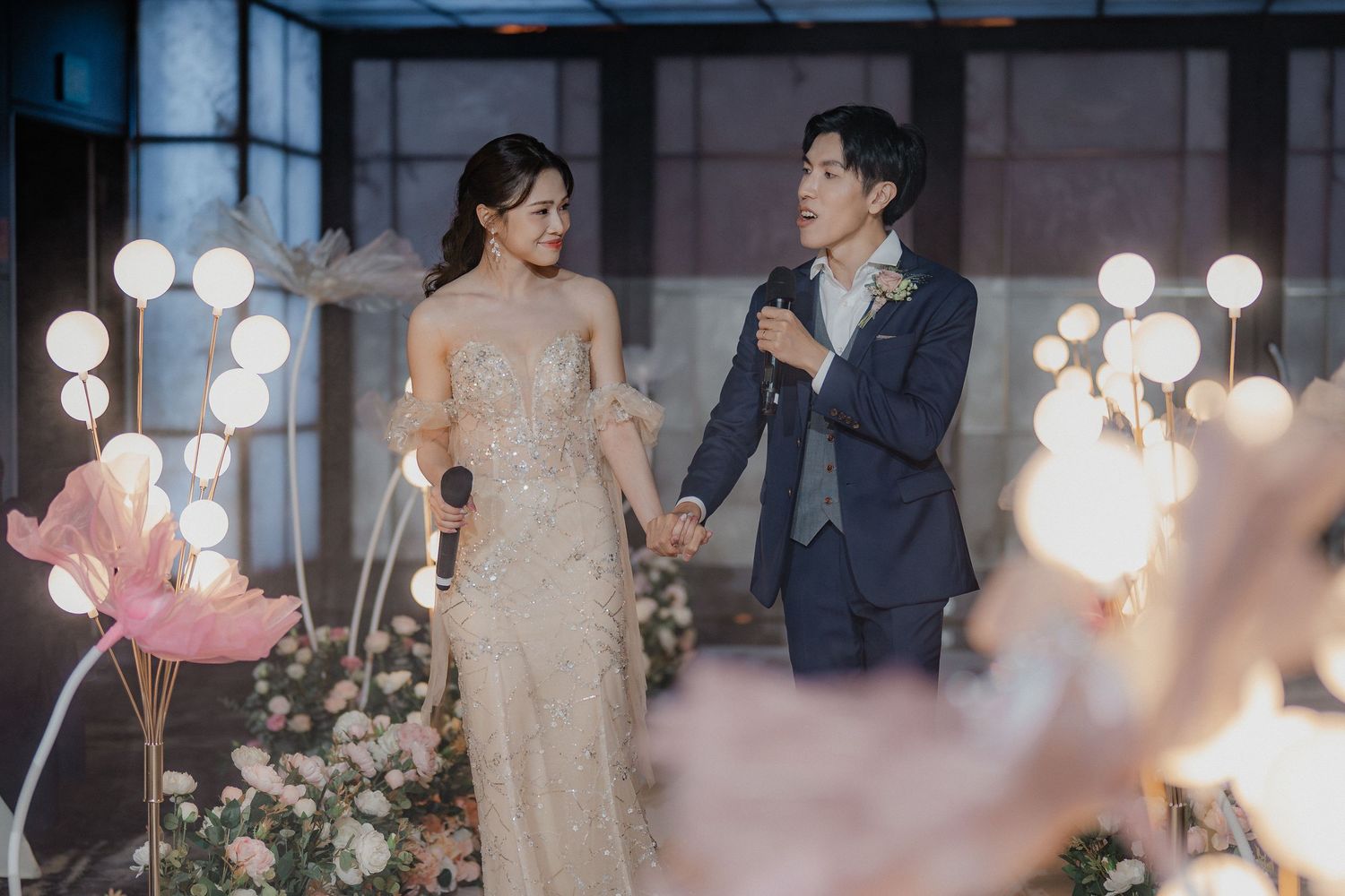 A couple in elegant attire pose together against a backdrop of glowing lights and floral decorations.