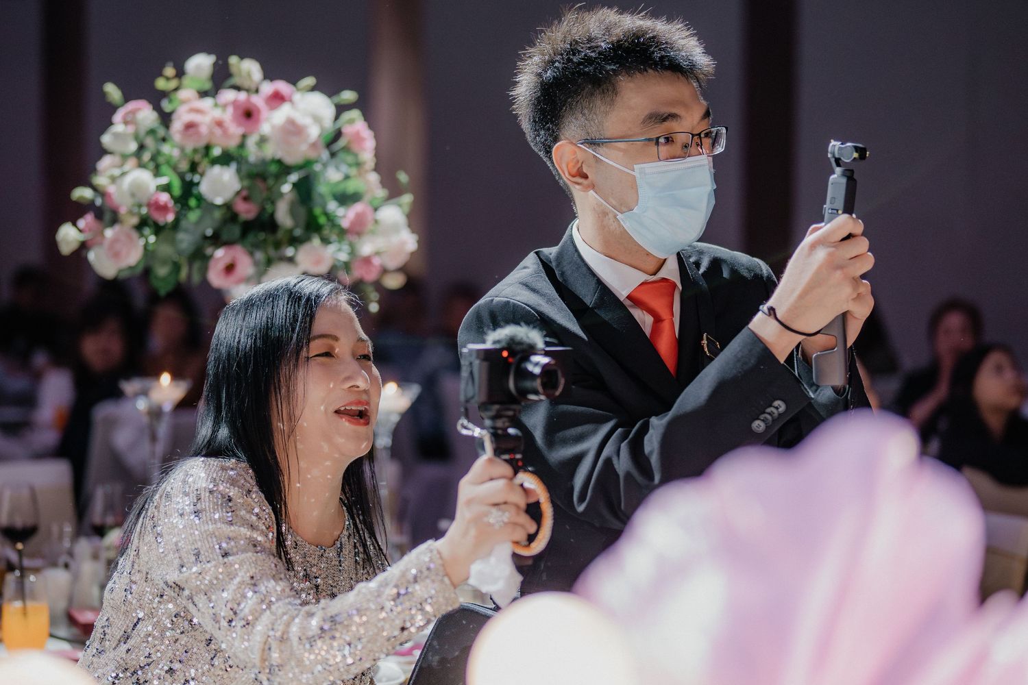 Wedding guests in formal attire hold up phones to capture moments during reception.