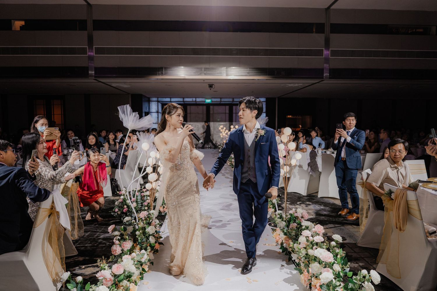 Newlyweds walk down aisle in modern indoor ceremony with floral decorations and seated guests.