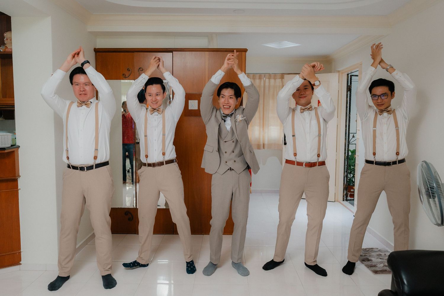 Groomsmen in beige suits striking fun poses against a white wall during wedding preparations.