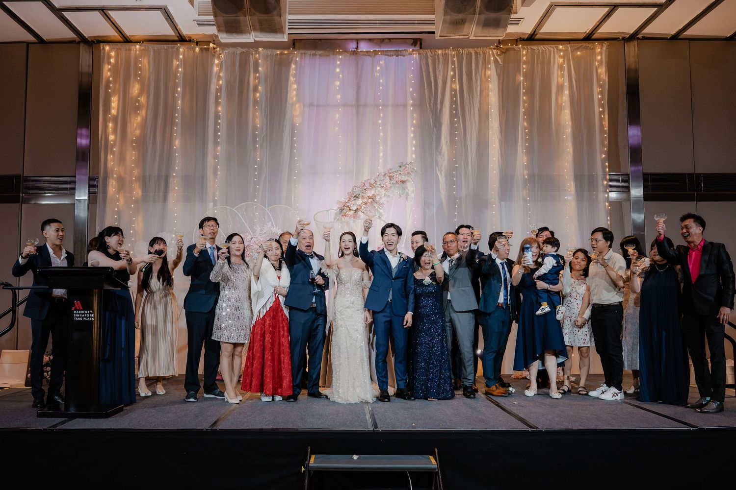 Large group wedding photo with guests posing in front of illuminated backdrop.