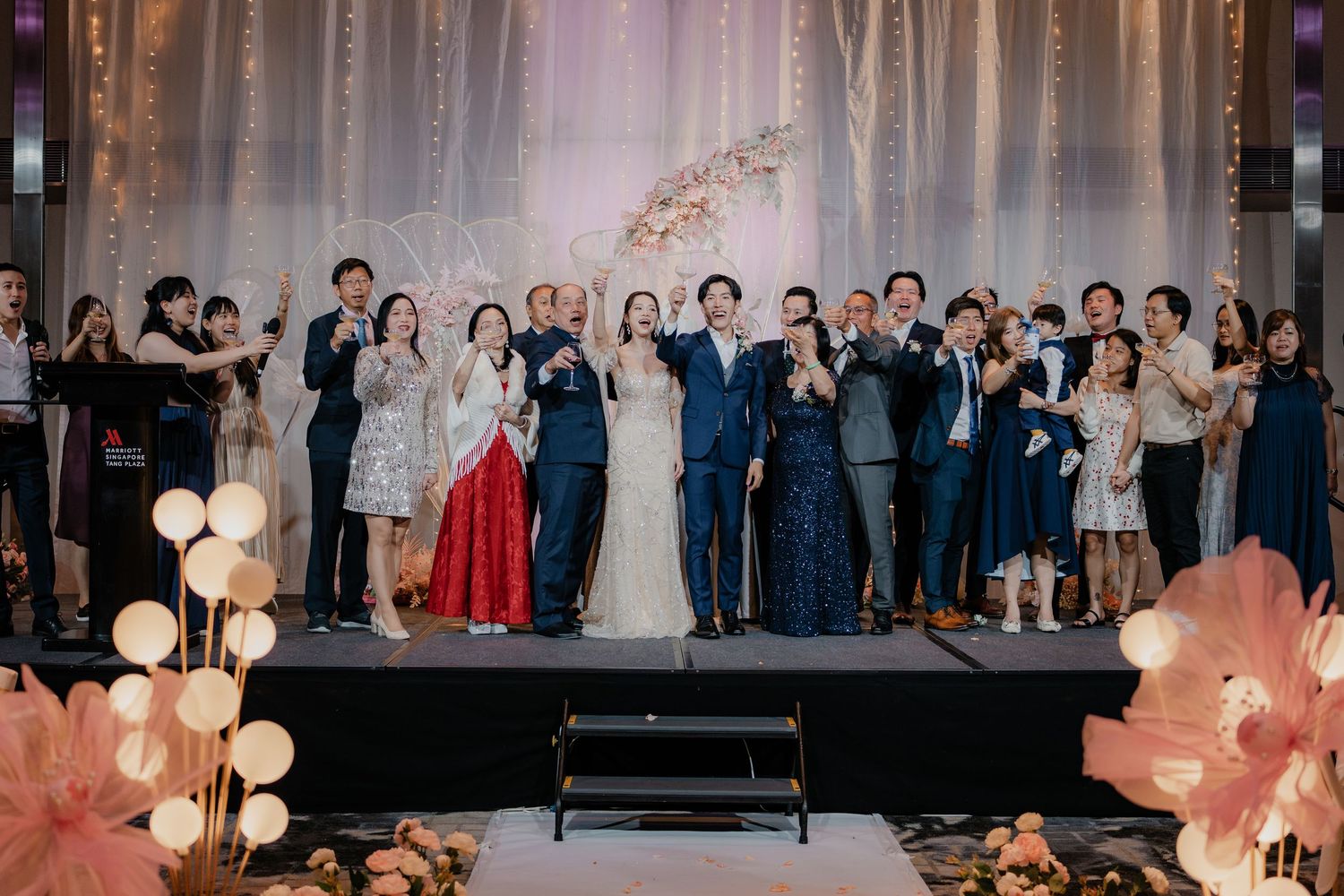 Wedding guests and party members raise glasses in celebration during an indoor reception with stage lighting.