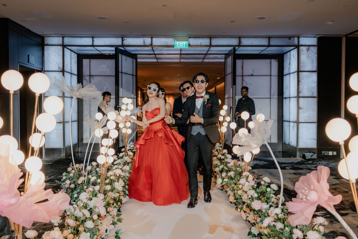 A couple makes their entrance through a hallway decorated with white balloons and pink floral arrangements.