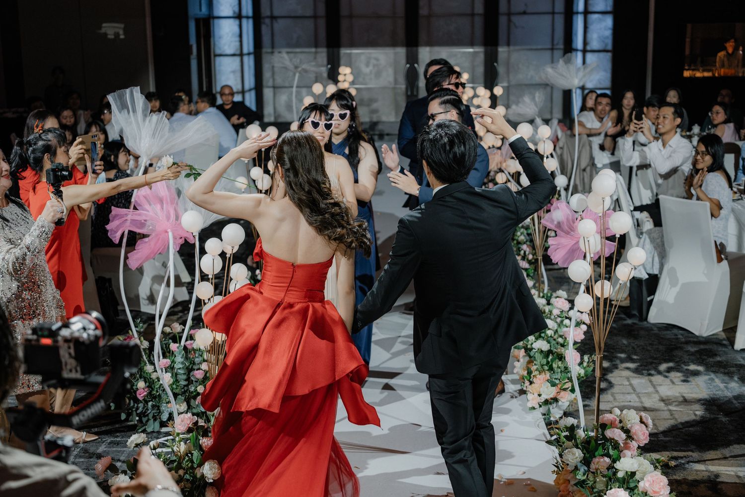 Wedding guests cheer and celebrate as couple dances beneath floral decor at an elegant indoor reception ceremony.