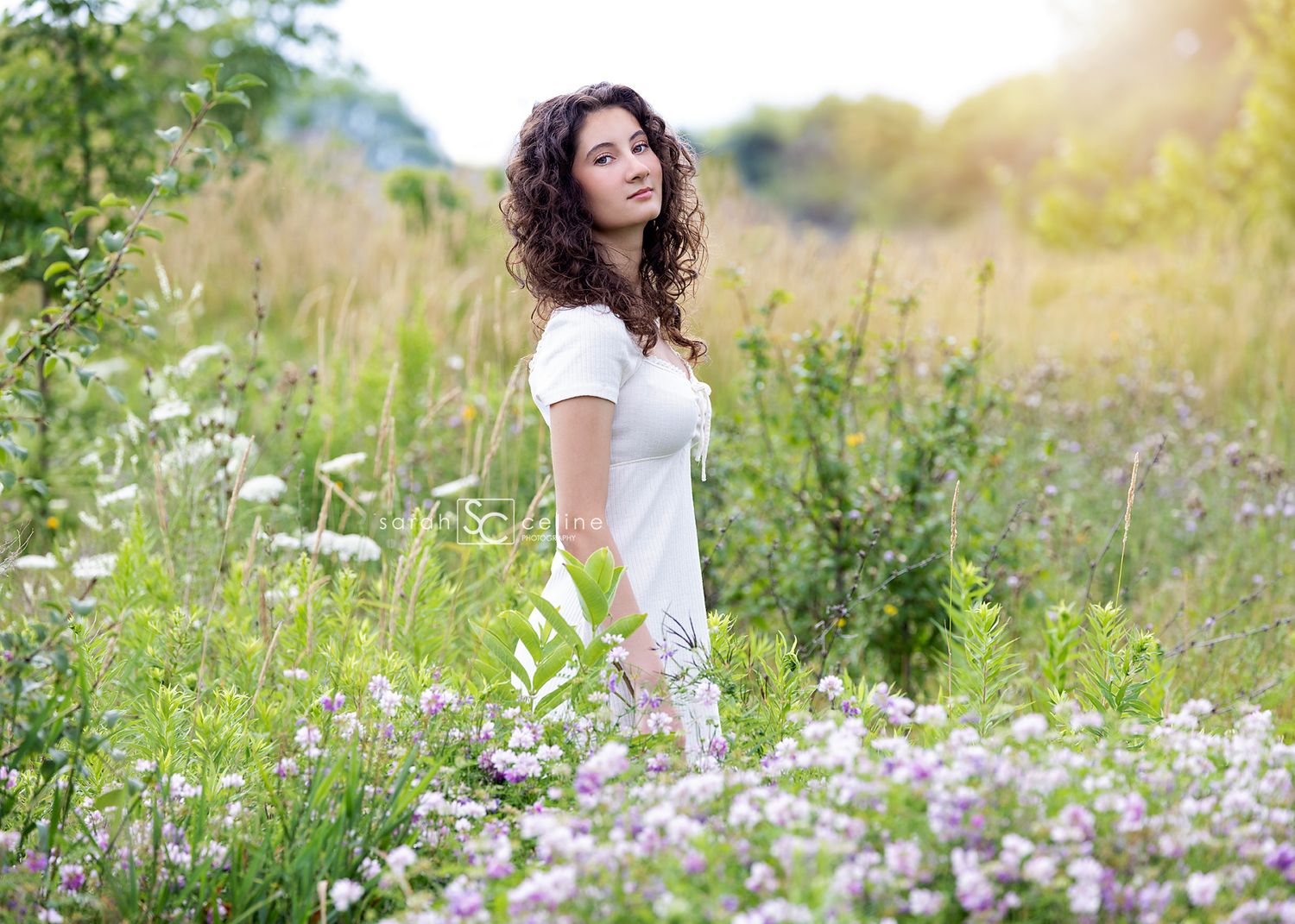 A person in a white dress stands in a wildflower meadow with tall grass and sunlit flowering plants.