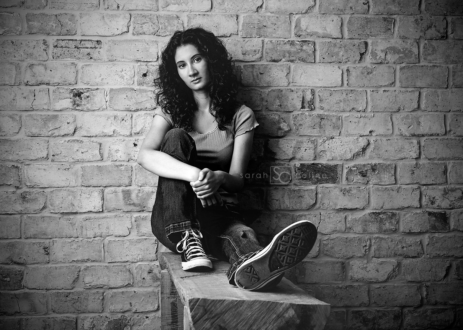 A black and white portrait shows a person sitting on a ledge against a brick wall wearing casual sneakers and jeans.