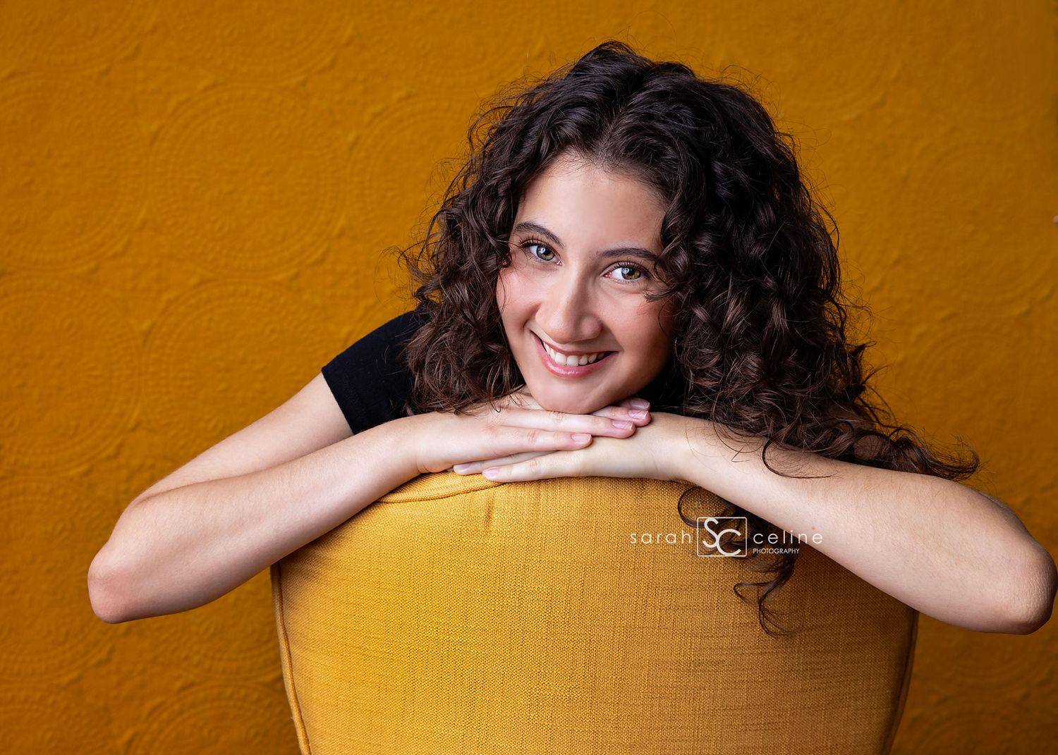A candid portrait of someone with dark curly hair leaning against a chair against a golden yellow wall.