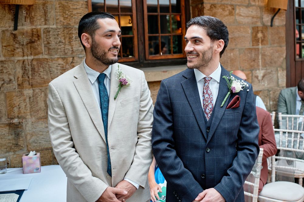Two people share a joyful moment at a wedding ceremony while wearing contrasting light and dark suits with boutonnieres.