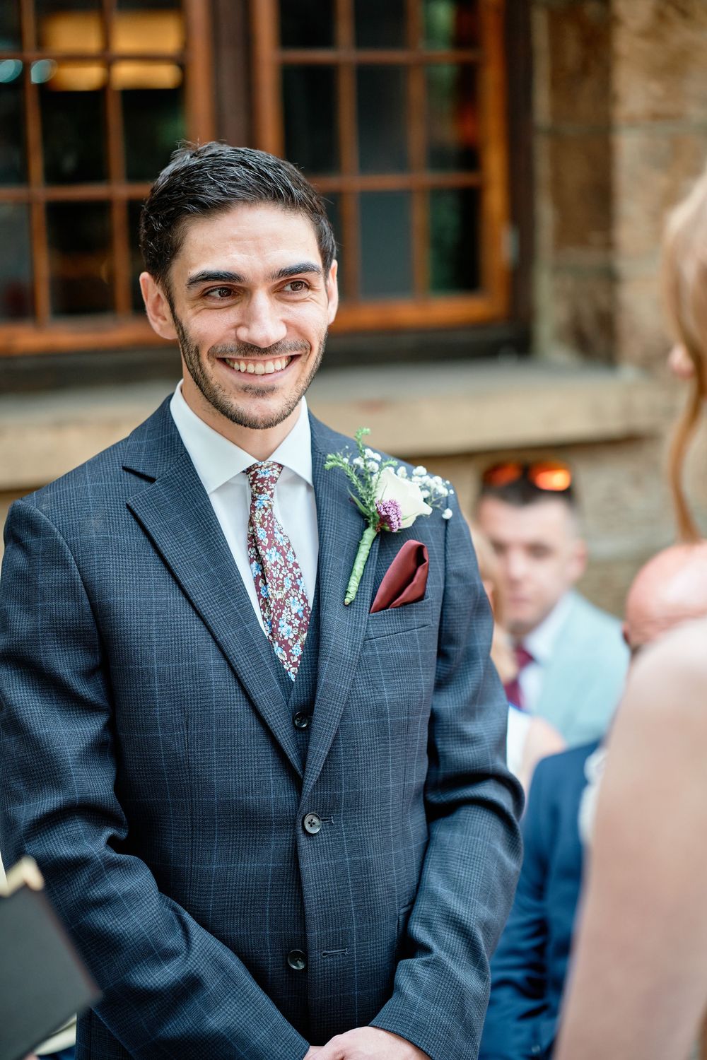 A groom in a navy suit with floral tie and burgundy pocket square smiles during his wedding ceremony.