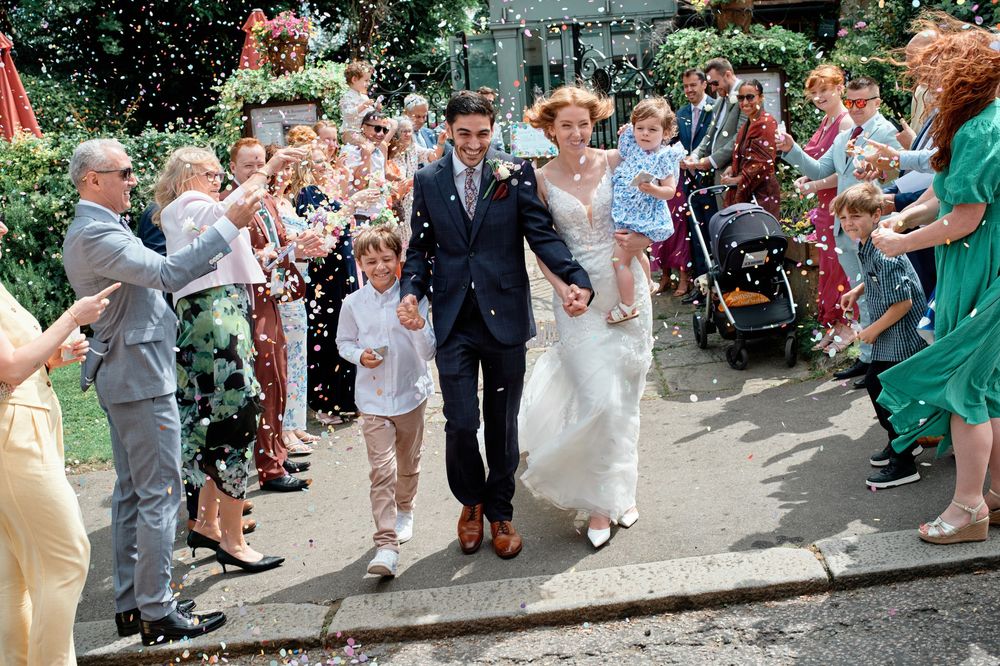 A crowd of wedding guests stands outside cheering and celebrating on a sunny day.