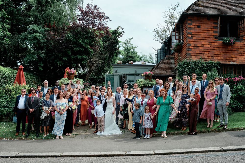 Large wedding group poses outdoors for photos on a bright summer day with a rustic brick building in the background.
