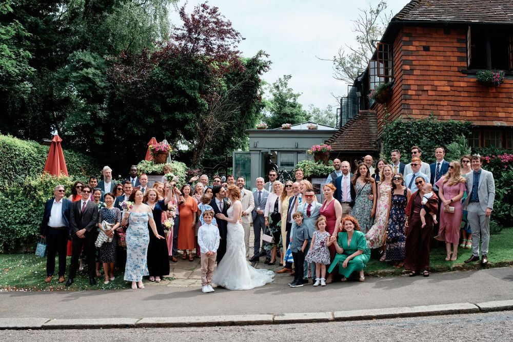Large wedding party gathers for a group photo outside a rustic red brick building on a sunny day.