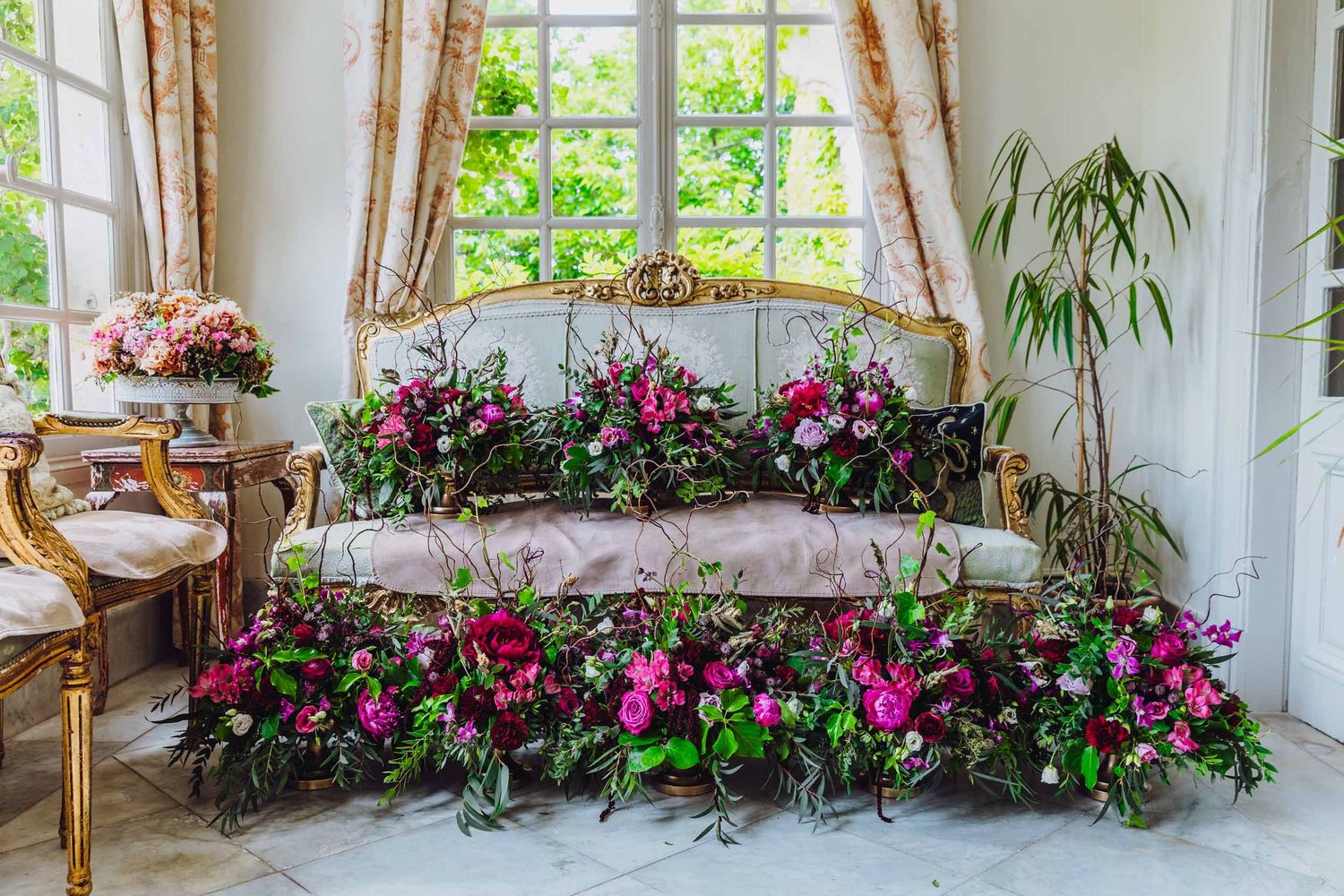 A sunny greenhouse interior features colorful floral arrangements, potted plants, and window-side seating for nature viewing.
