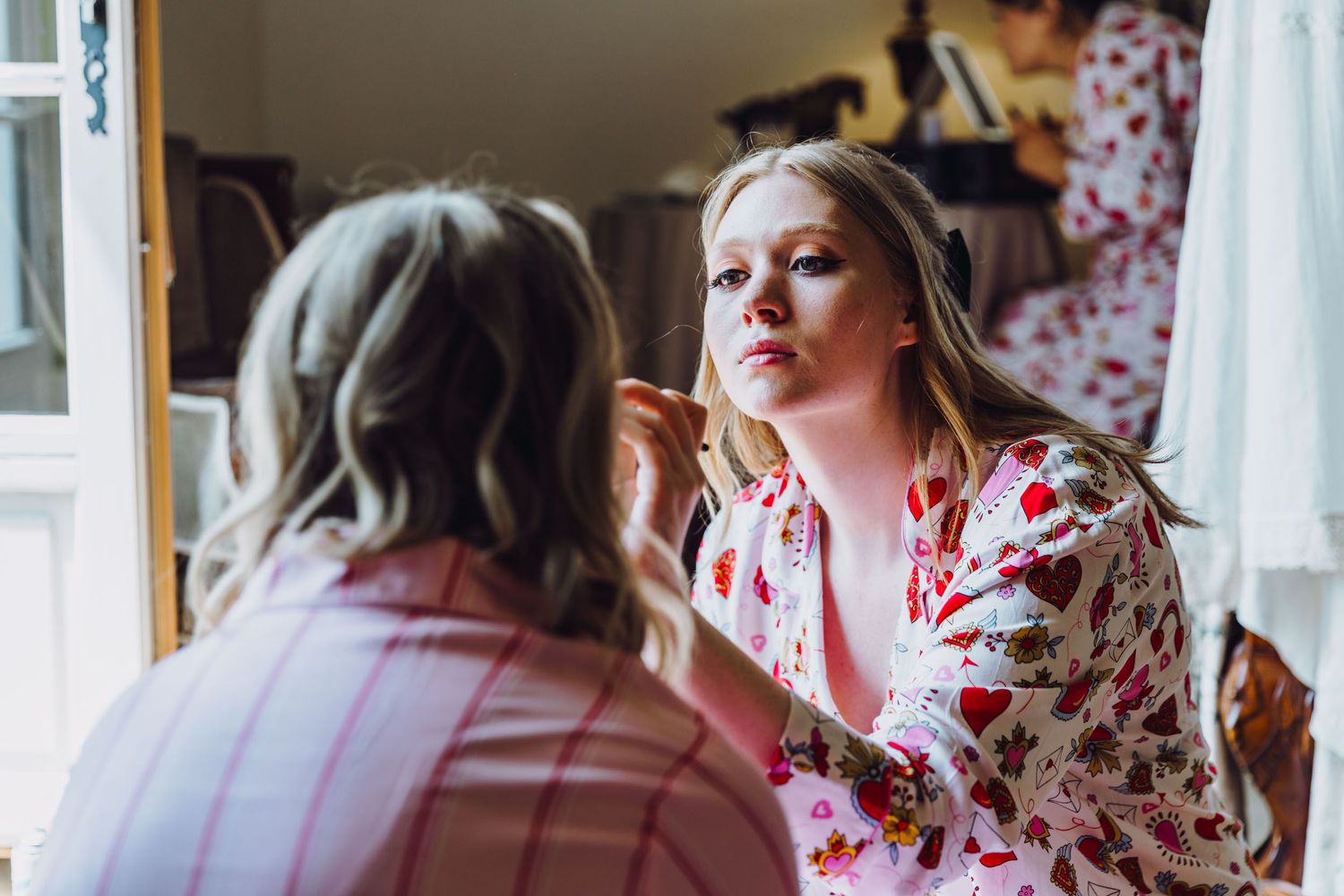 Someone applying makeup in a kimono-style floral robe while sitting at a vanity mirror in natural light.