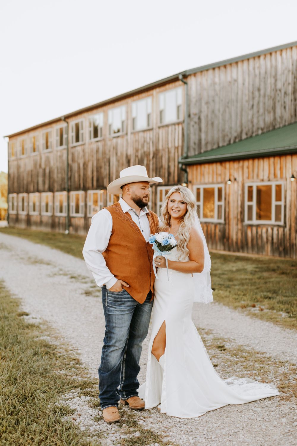 A rustic country wedding portrait in front of a weathered wooden barn with a cowboy theme and warm autumn colors.
