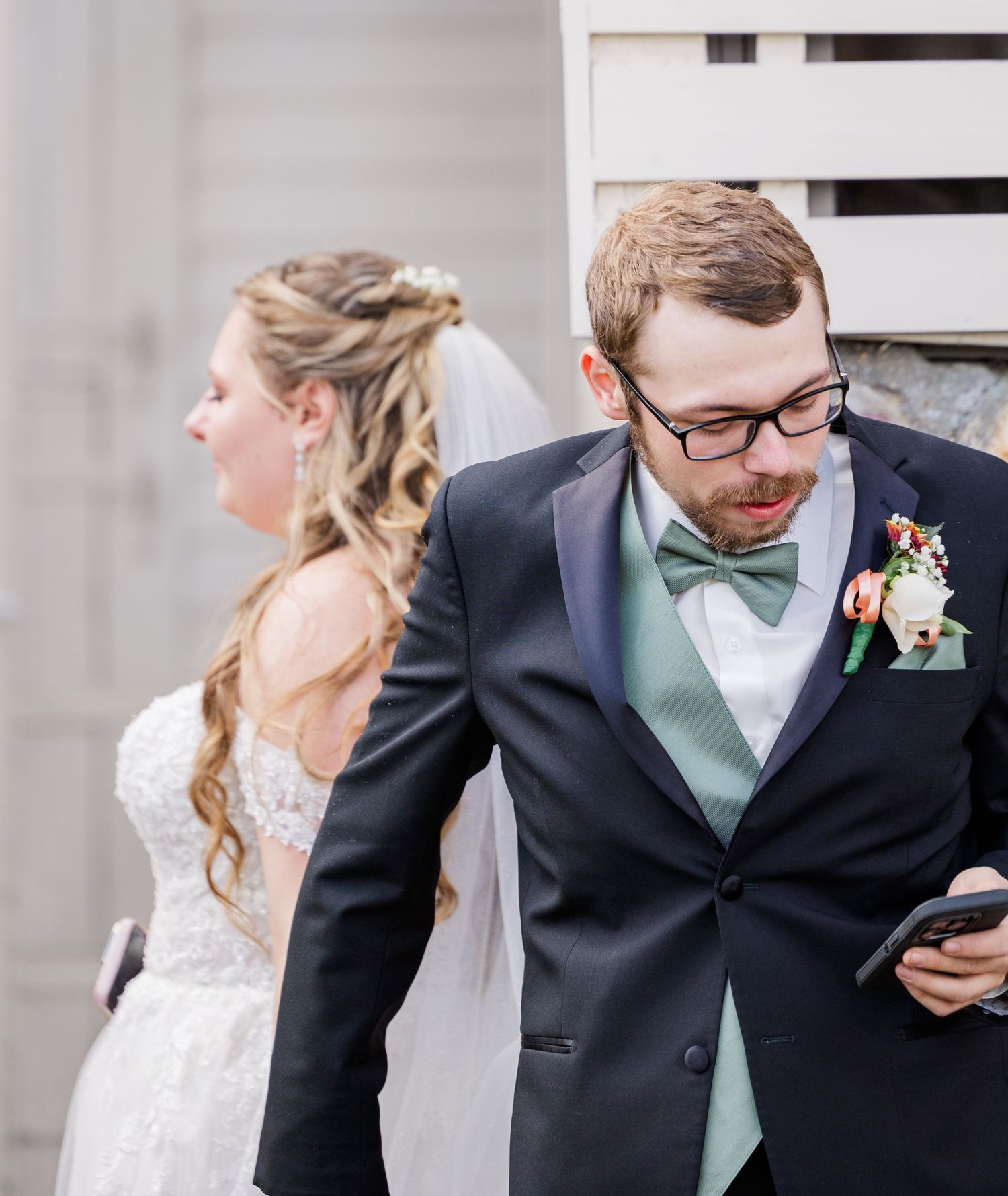 A bride and groom stand back-to-back while looking at their phones during their wedding.