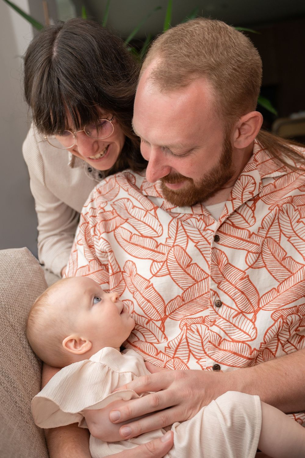 À Saint-Hubert, séance photo famille à domicile d’un couple assis dans un fauteuil tenant leur bébé dans les bras, regard complice et moment