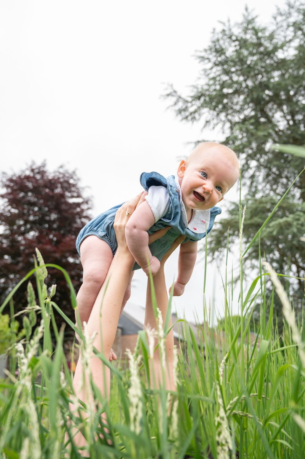 À Saint-Hubert, séance photo bébé en extérieur, bébé porté en l’air dans l’herbe regardant vers la caméra