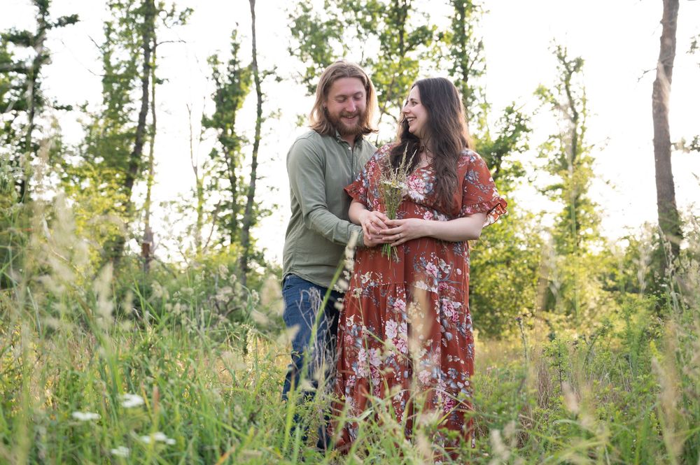 Couple attendant un bébé à Barvaux riant naturellement et tenant un bouquet de fleurs lors d’une séance photo grossesse