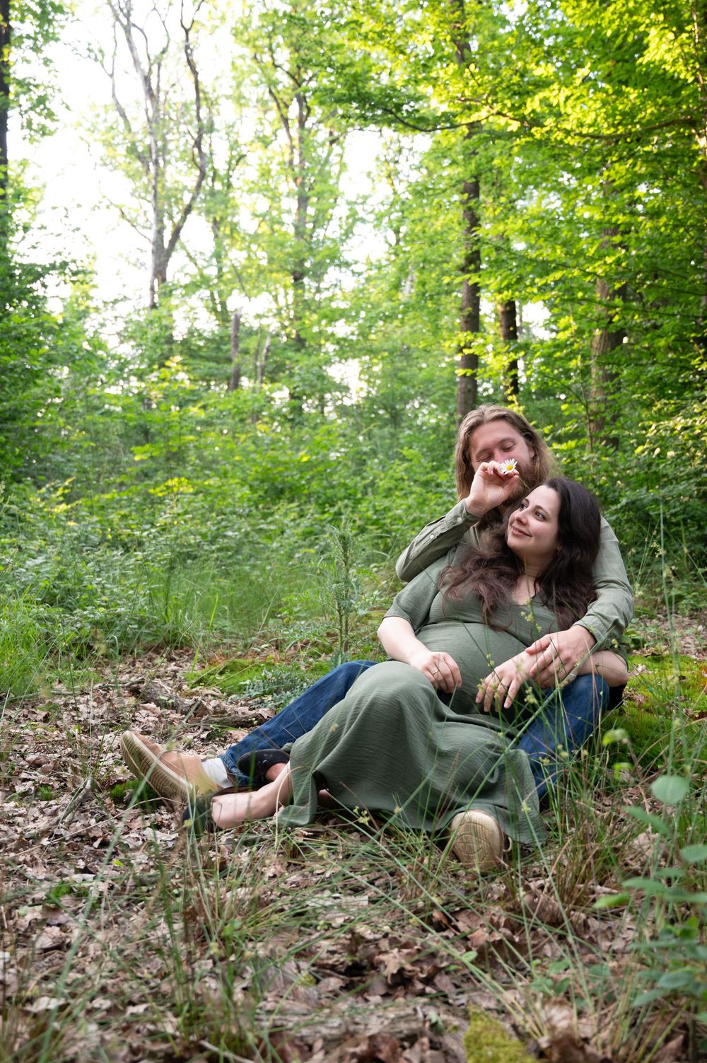 Couple allongé en forêt à Barvaux lors d’une séance photo grossesse, le papa déposant une fleur sur l’oreille de la maman