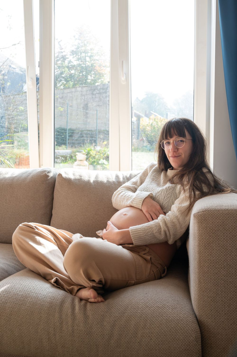 À Saint-Hubert, future maman photographiée à domicile assise dans un fauteuil devant une baie vitrée lors d’une séance photo grossesse