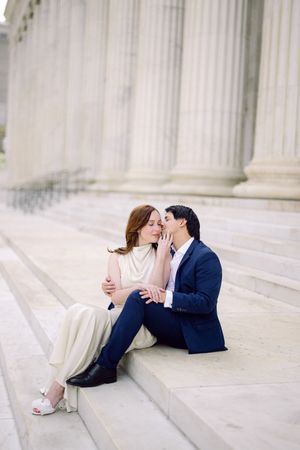 A romantic couple sits on white marble steps outside a building with classical columns in the background.