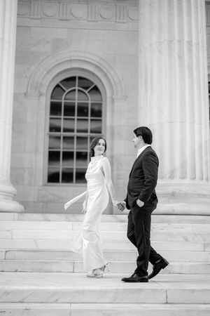 Elegant couple walking up stone steps of a classical building with ornate arched window and columns in black and white.