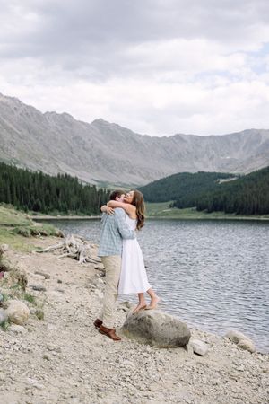 Couple embracing on rocky shore of mountain lake surrounded by pine trees and peaks in Colorado.