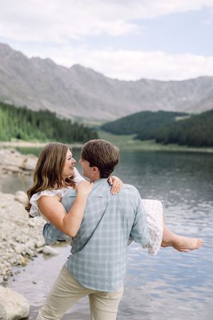 Man carrying woman in his arms by a mountain lake with forested peaks reflected in calm water.