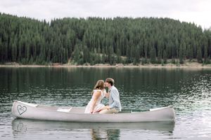 Couple sharing a romantic kiss in a canoe on a serene mountain lake surrounded by evergreen forests.