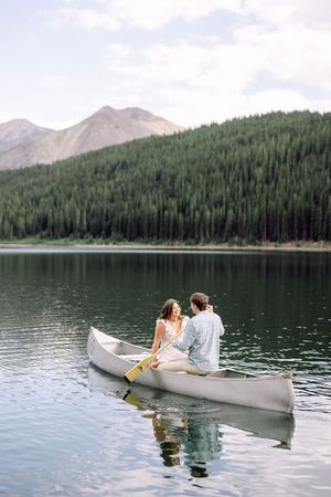 Couple sitting in a white canoe on a calm mountain lake surrounded by pine trees and scenic peaks.