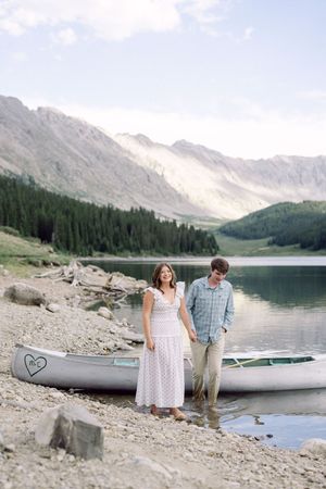 Couple standing by a canoe on a rocky mountain lake shore, surrounded by pine trees and scenic peaks.