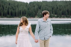 Couple holding hands by a mountain lake, woman in white dress and man in plaid shirt, with pine trees in background.