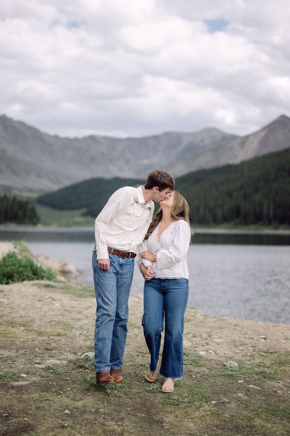 Couple kissing near a mountain lake in Colorado, both wearing white tops and jeans, with scenic peaks and clouds behind them.