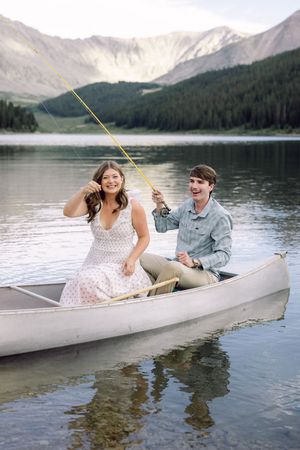 Couple laughing in a canoe while fishing on a serene mountain lake surrounded by pine forests.