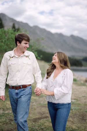 Couple in white shirts and jeans holding hands, smiling at each other with mountain lake backdrop.
