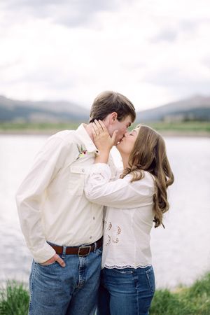 Couple sharing a romantic kiss by a mountain lake, both wearing white shirts and jeans in an outdoor engagement photo.