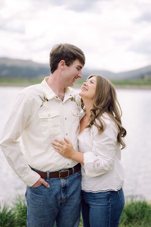 Couple smiling at each other in white shirts and jeans near a mountain lake during an outdoor engagement photo session.