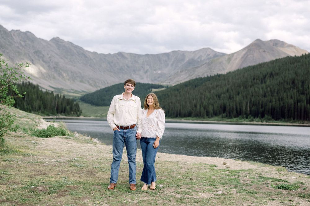 Happy couple holding hands and smiling before a scenic mountain lake surrounded by pine forests.