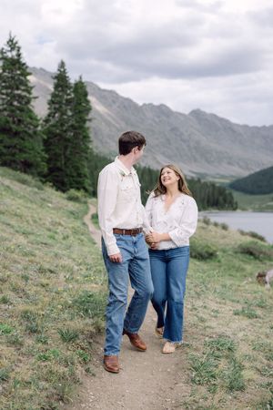 Couple walks hand-in-hand on mountain path near alpine lake, wearing white shirts and jeans, smiling at each other.