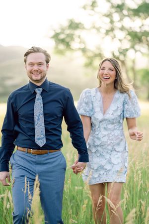 A couple holds hands while walking through a sunlit meadow, wearing blue formal attire and a floral dress.