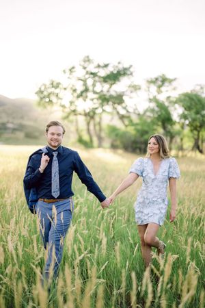 A playful couple runs and chases through a tall grass field at sunset wearing casual clothing.