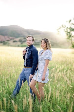 A couple walks hand in hand through a sunlit meadow of tall grass with mountains in the background at sunset.